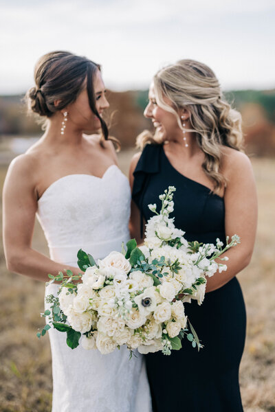 woman in white dress hugging and smiling at woman in black dress while holding bouquets 