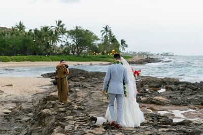 A photographer taking a picture of a newlywed couple standing on a rocky shoreline