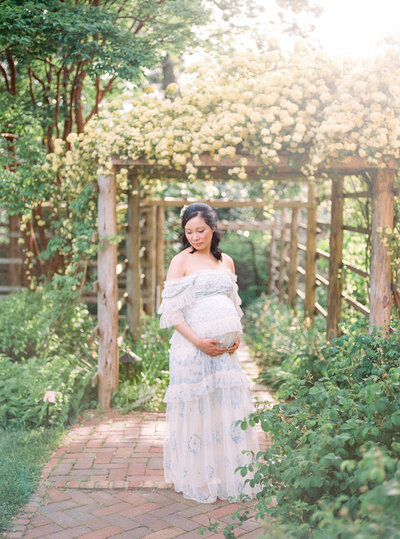 A pregnant mother in a blue dress standing under a pergola of white flowers