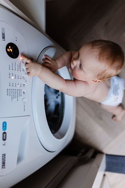 Foto van bovenaf baby staat in pamper bij wasmachine en drukt op een knopje
