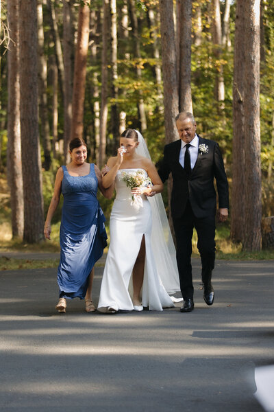 Bride walks down the aisle with her arms intertwined with both her mother and father. She is dressed in a tight fitting satin wedding dress with a slit up her left leg. She has a cathedral length wedding veil in. She wipes a tear with a tissue in one hand and she has her light pink and white floral bouquet in the other. They are surrounded by the tall pine trees at Catalyst by Nature Link.