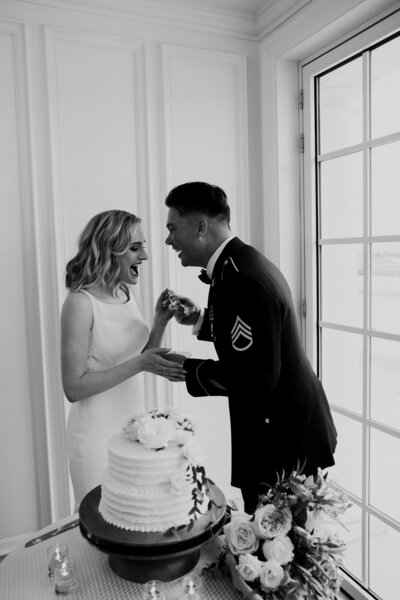 Groom in uniform and bride in elegant dress playfully fight about the cake feeding