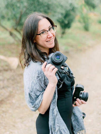 bay area photographers posing for a head shot as she holds her cameras in both hands and looks over her shoulder and smiles on an outdoor trail in Marin County
