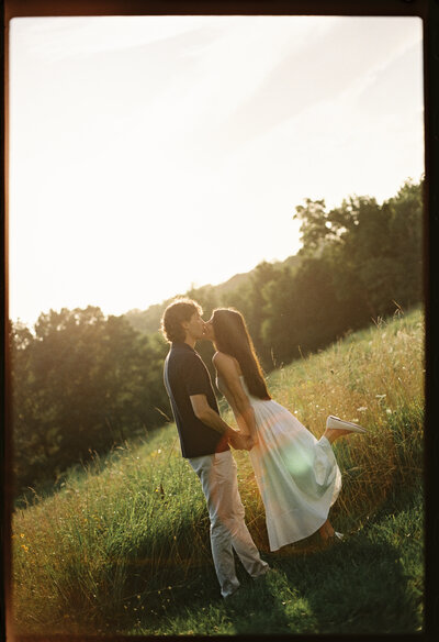 A 35mm film photo of a couple kissing in a grassy field at golden hour with the sun flaring behind them.