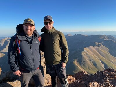 two men in baseball camps and hiking clothes on top of a mountain with views of range below