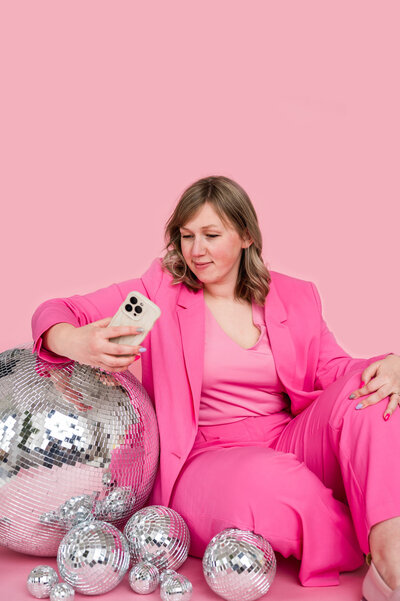 Kat Murphy in a pink suit sitting with her arm on a disco ball holding her phone in front of a pink background