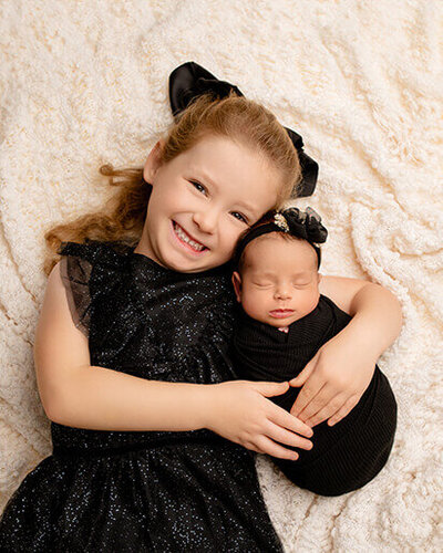 Proudn big sister in a black dress smiling big as she holder her new baby sister, who is also wearing black.