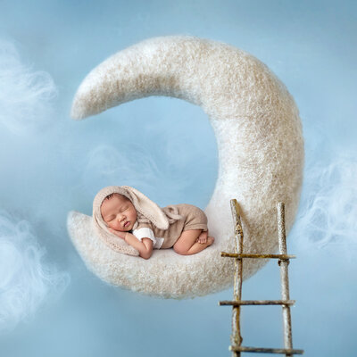 Newborn baby boy sleeping in a crescent moon prop with a soft blue sky backdrop and fluffy white clouds.