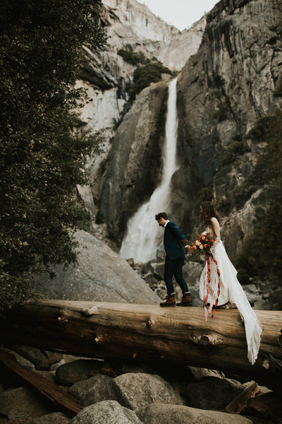 bride and groom walking on log over water with waterfall in background
