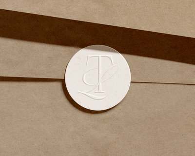 A white circular seal with embossed overlapping letters "T" and "L" is placed on a brown paper surface with diagonal shadows, reflecting Sweet Pea Ink Creative’s attention to detail in brand design.