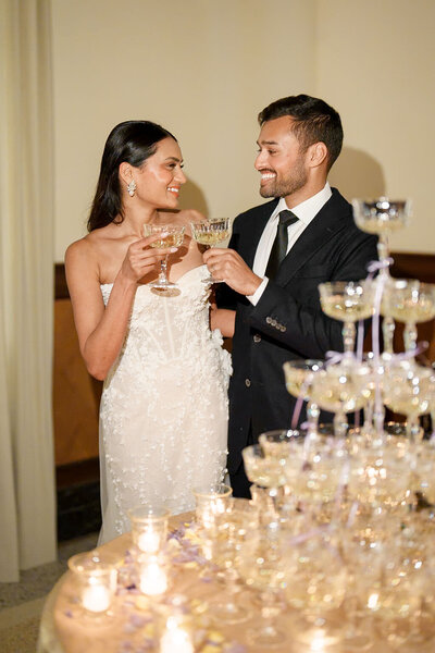 bride and groom smiling at each other while standing in front of champagne tower at wedding reception