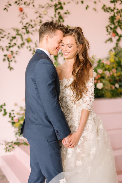 A bride and groom face-to-face while their hands are interlocked standing in front of a floral pink wall found inside of The Doyle in Las Vegas, Nevada.