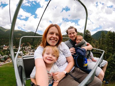 Picture of a family sitting on a white cable car. The woman is wearing a white t-shirt, the child is smiling, and the man is carrying the baby and a blue luggage