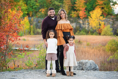 Mom and dad with their two daughters standing on a rocky cliff in front of fall foliage for their fall family photographer session in Burlington, Ontario.