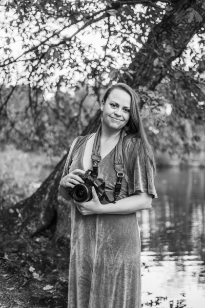 Newborn photographer Lindsay Wilson stands with her camera outside and smiles at camera.