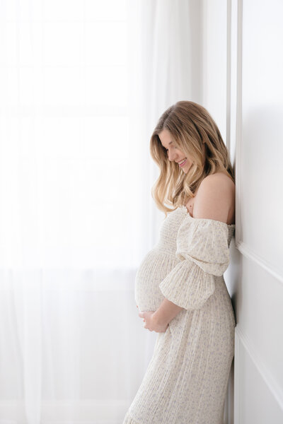 Expecting mom leaning against a wall dressed in a beautiful floral gown smiling and looking down at her bump