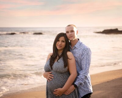 Expectant mom and dad embracing baby bump on California beach with pink skies at holden hour.