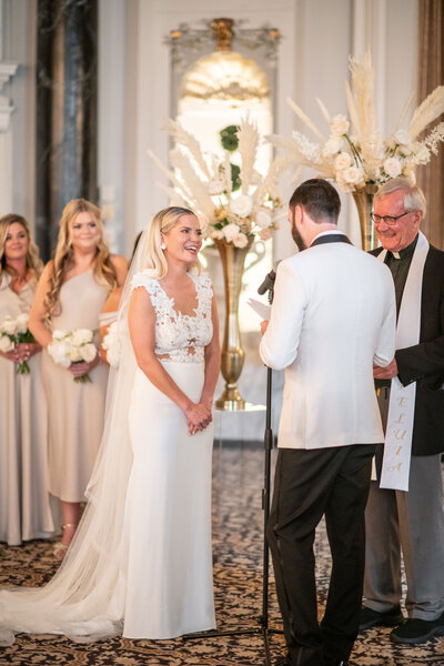 A bride and groom stand facing each other during their wedding ceremony. The bride is smiling, wearing a white gown and veil, while the groom is in a white suit jacket. Bridesmaids in matching dresses stand nearby, and a clergyman officiates the ceremony.