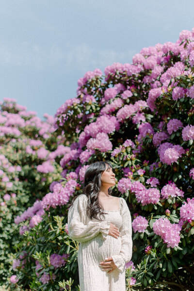 A pregnant woman is holding her belly and looking up to the sky with a big rhododendron bush behind her blooming