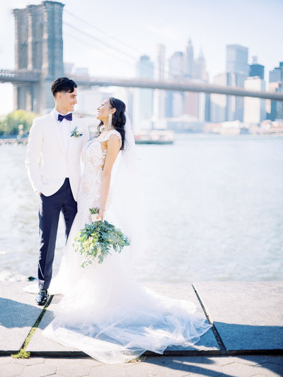 Bride and groom looking at each other fondly as they stand over the Brooklyn Bridge at Pebble Beach in DUMBO during their New York City wedding captured by Tom Schelling Photography.