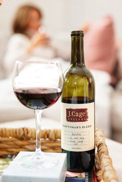 Glass of wine and wine bottle with woman drinking wine in background.