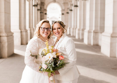 Two woman one dressed in a white suit and the other in a wedding dress hold a bouquet and smile at Union Station in Washington DC