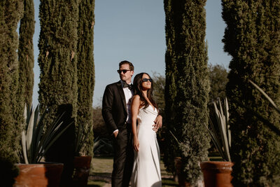 Stylish bride and groom standing between tall cypress trees. The woman wears sunglasses and a white gown, while the man stands beside her in a classic black suit, exuding confidence and cinematic elegance.