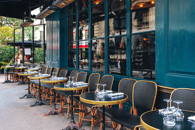 An outdoor café with small round tables and woven bistro chairs arranged in rows along a teal-colored storefront, each table set with upside-down wine glasses; the street scene appears quiet with no patrons seated.
