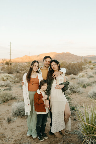 A family of four, with two adults and two children, posing together in a desert landscape during sunset. they are smiling and dressed in casual, layered clothing.