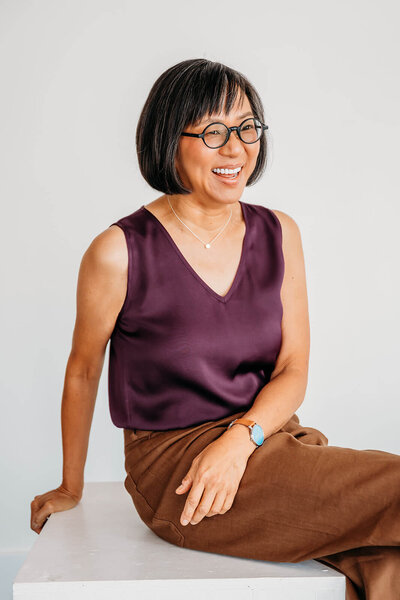Smiling woman with short hair, wearing a purple sleeveless top, seated with hands clasped.