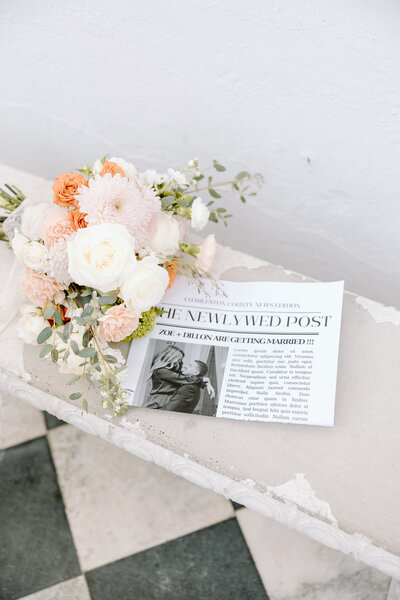Romantic Charleston wedding flatlay featuring a pastel bridal bouquet and a personalized newspaper-style announcement on a vintage bench.