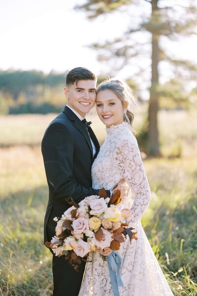 Portrait of a handsome young man with dark brown hair in a black tux with bow tie next to a smiling woman in a wedding dress with a big bouquet of fall blooms at Bluebird Meadows Farm in Raleigh.