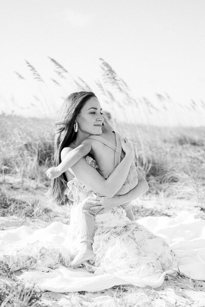 A mom hugging her baby on the beach while the wind blows their hair