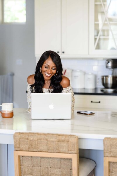 Deme Riley working at a kitchen table with her laptop, sharing journal prompts to boost personal transformation.