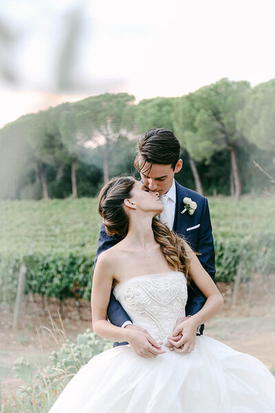 Tuscany backdrop with vineyard for a newlyweds photo. Groom is kissing the bride's forehead. Luxury wedding gown and tuxedo. 