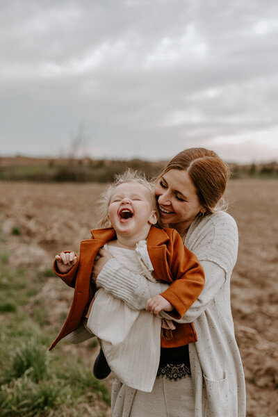 Mom and toddler in Exeter, Ontario field for family photoshoot. Mom holding toddler so she is facing the camera. Toddler is laughing and mom is snuggling her with her cheek against the little girl's head.