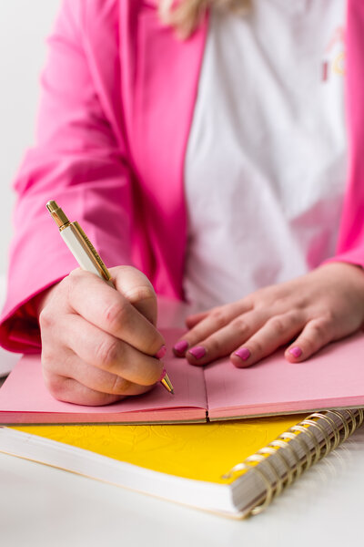 Hand with pink fingernails writing in a pink notebook with a gold pen