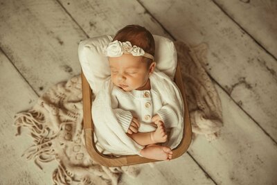 Precious moments of your newborn baby girl with our heartwarming photography session. Watch as she peacefully sleeps in a small and cozy basket, wearing a sweet white outfit.