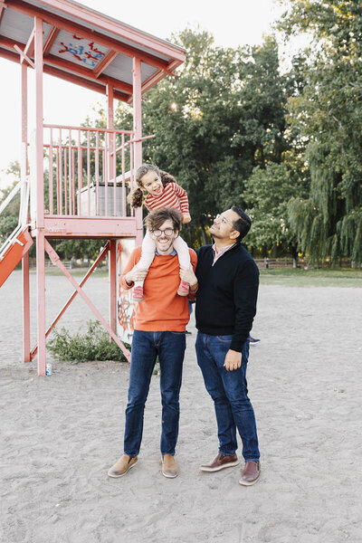 family with 2 dads and toddler girl having fun at Cherry Beach. LIttle girl is on dads shoulders.