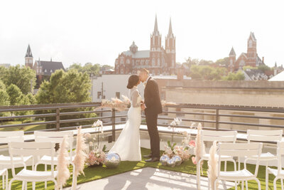 bride and groom standing outside with trees and large buildings in the background while kissing
