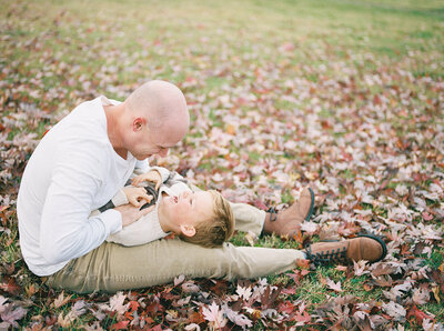 Dad, sitting in a pile of leaves, tickles 3 year old son as he giggles taken by Little Rock film photographer Bailey Feeler