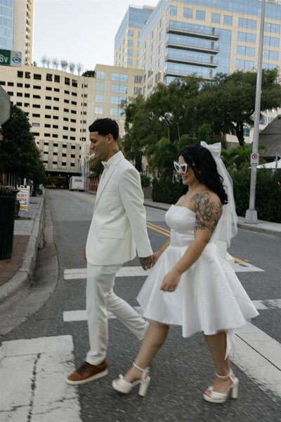 Bride and groom in white outfits walk hand in hand across a downtown street during their Orlando elopement, with city buildings in the background.