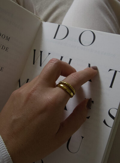 Hand wearing gold rings resting on an open book, styled detail for The Maddison Edit brand.