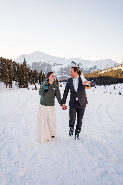 A bride and groom smile at each other while holding hands and walking towards the camera carrying skis