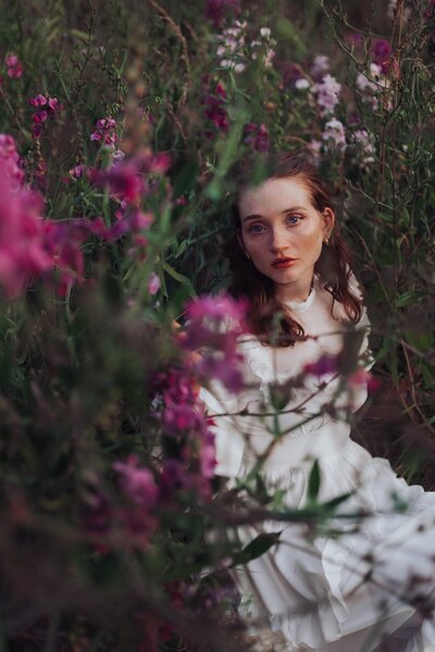 Young, red-headed woman sits in a flowy white dress in a field of flowers