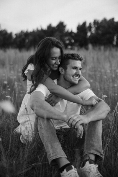 Woman drapes her arms around her fiance's shoulders as he smiles off into the sunset
