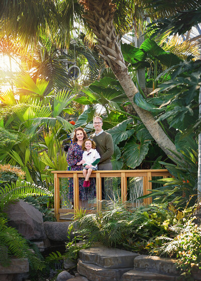 smiling family posed on a small bridge in a conservatory with tropical plants and trees surrounding them