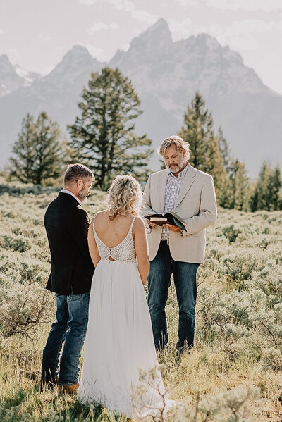 grand tetons in the far distance with trees closer to the bride and groom as they say their vows together in the meadow in front of the forest captured by jackson hole wedding photorgaphers