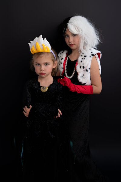 Two young girls standing in front of black backdrop, dressed in Ursula and Cruella costumes with evil looks on their faces.