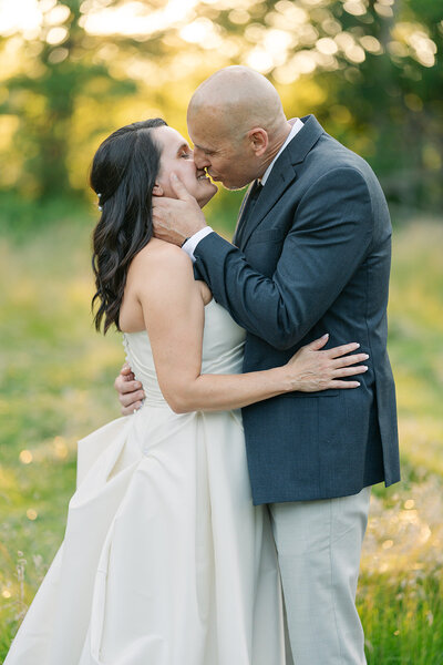 Newlyweds embracing one another and kissing outside in front of trees at sunset. 